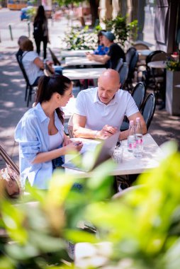  İnsanlar dışarıda bir toplantıda konuşuyorlar. Esmer kadın ve orta yaşlı adam kaldırımdaki kafede oturuyor. 