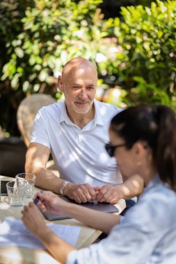  İnsanlar dışarıda bir toplantıda konuşuyorlar. Esmer kadın ve orta yaşlı adam kaldırımdaki kafede oturuyor. 