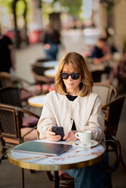 Orta yaşlı bir kadın kaldırımdaki kafede otururken mesaj atıyor. Sarı saçlı, güneş gözlüğü takan ve günlük kıyafetler giyen bir kadın..  