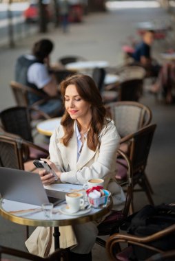 Masada oturan neşeli bir kadın, açık bir kafede akıllı telefon kullanıyor. Trençkot giyen çekici bir kadın.. 
