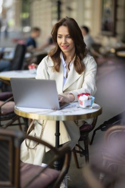 Masada oturmuş dışarıda bir kafede dizüstü bilgisayarla çalışan neşeli bir kadın. Trençkot giyen çekici bir kadın. Kendine güvenen profesyonel kadın video görüşmesi yapıyor..  