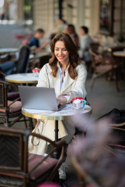 Masada oturmuş dışarıda bir kafede dizüstü bilgisayarla çalışan neşeli bir kadın. Trençkot giyen çekici bir kadın. Kendine güvenen profesyonel kadın video görüşmesi yapıyor..  