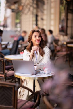 Masada oturmuş dışarıda bir kafede dizüstü bilgisayarla çalışan neşeli bir kadın. Trençkot giyen çekici bir kadın. Açık havada çalışan kendine güvenen profesyonel bir kadın..  