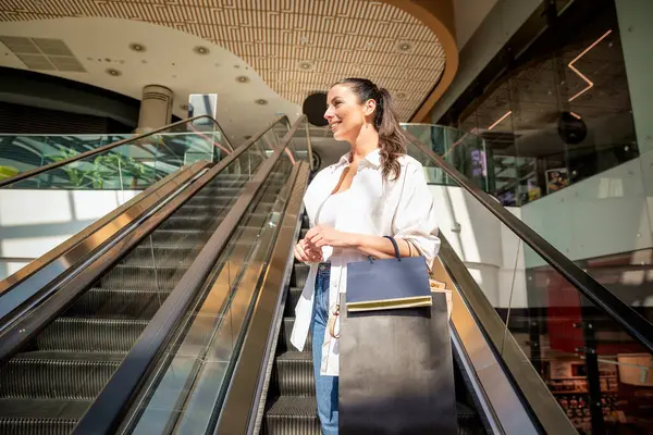 Esmer kadın yerel bir alışveriş merkezinde yürüyen merdivenle aşağı iniyor. Alışveriş torbaları taşıyor ve aldıkları kıyafetlerle mutlu..