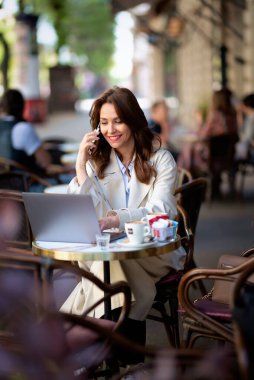 Masada oturan neşeli bir kadın, dizüstü bilgisayarla çalışıyor ve açık bir kafede akıllı telefon kullanıyor. Trençkot giyen çekici bir kadın..  