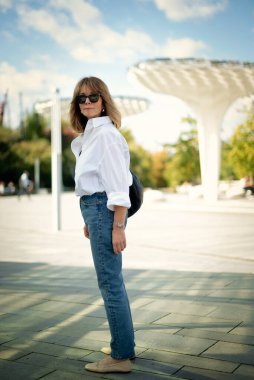 Şehir parkında yürüyen orta yaşlı bir kadın. Beyaz gömlek ve kot giyen sarışın kadın. Tam boy çekim. 