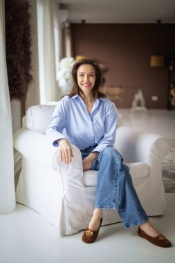 Güzel bir kadın evde rahatlatıcı bir günün tadını çıkarıyor. Orta yaşlı kadın rahat koltuğunda otururken ışıl ışıl gülümsüyor. Tam boy çekim.  