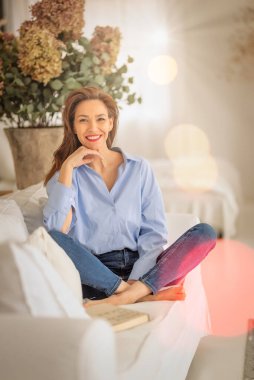 Güzel bir kadın evde rahatlatıcı bir günün tadını çıkarıyor. Orta yaşlı bir kadın, rahat kanepesinde uzanmış kitap okurken ışıl ışıl gülümserdi.. 