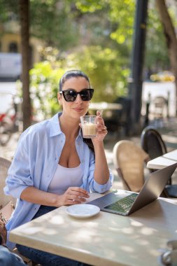 Masada oturmuş gülümseyen bir kadın, açık bir kafede dizüstü bilgisayarla çalışıyor. Sıradan kıyafetler giyen çekici bir kadın. Kendine güvenen profesyonel kadın video görüşmesi yapıyor..  