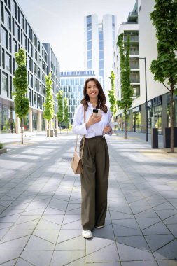 İş yerindeki ofise yürüyen kendine güvenen bir iş kadını. Telefonunu ve mesajlarını kullanan profesyonel bir kadın. Esmer, iş kıyafeti giyen bir kadın. Tam boy çekim.  