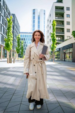 Orta yaşlı bir iş kadınının elinde dizüstü bilgisayarla iş bölgesinde yürümesi. Çekici profesyonel kadın iş kıyafeti giyiyor ve neşeli gülümsüyor.. 
