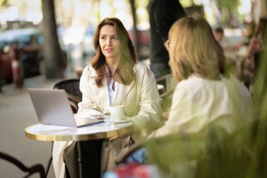 İki orta yaşlı kadın bir kafede terasta oturuyor ve konuşuyorlar. İki bayan arkadaş gündelik kıyafetler giyip kahve içiyorlar..