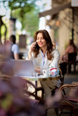 Masada oturan neşeli bir kadın, dizüstü bilgisayarla çalışıyor ve açık bir kafede akıllı telefon kullanıyor. Trençkot giyen çekici bir kadın..  