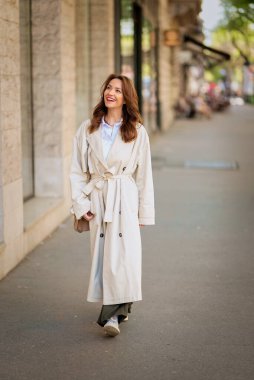 Güneşli bir günde ve vitrinlere bakan, uzun saçlı, kendinden emin, olgun bir kadın. Trençkot giyen ve neşeli gülümseyen çekici bir kadın. Tam boy çekim.  
