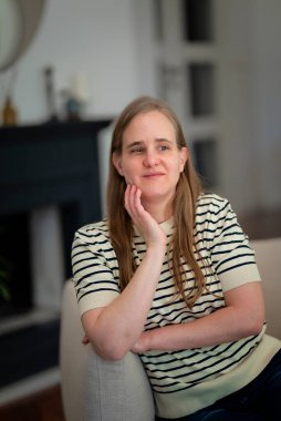 Görme engelli bir kadın evinde koltukta oturuyor. Sarı saçlı, sıradan kıyafetler giyen bir kadın..  