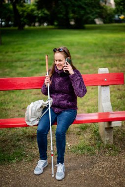 Beyaz baston tutan ve parktaki bankta oturan görme engelli bir kadın. Gülümseyen bir kadın telefonda konuşuyor ve günlük kıyafetler giyiyor. Tam uzunluk.  
