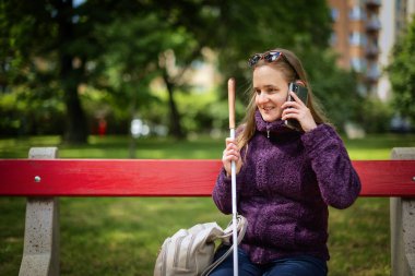 Beyaz baston tutan ve parktaki bankta oturan görme engelli bir kadın. Gülümseyen kadın rahat ceket ve kot pantolon giyiyor.. 