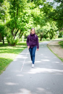 Beyaz bastonuyla parkta yürüyen görme engelli bir kadın. Sarı saçlı, sıradan kıyafetler giyen bir kadın. Tam boy çekim. 