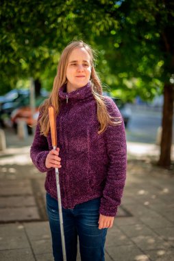 Beyaz baston tutan ve parkta duran görme engelli bir kadın. Gülümseyen kadın rahat ceket ve kot pantolon giyiyor..  