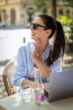 Masada oturmuş dışarıda bir kafede dizüstü bilgisayar kullanan neşeli bir kadın. Sıradan kıyafetler giyen çekici bir kadın..  