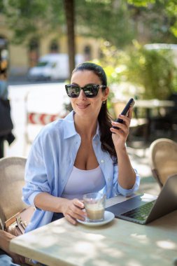 Esmer bir kadın masada oturuyor, dizüstü bilgisayarla çalışıyor ve açık bir kafede akıllı telefon kullanıyor. Gündelik kıyafetler ve güneş gözlüğü takan çekici bir kadın..  