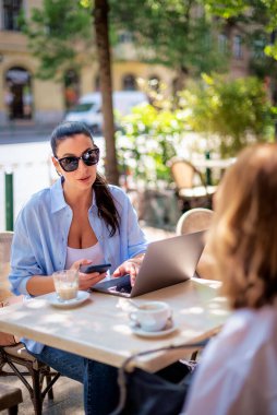 Esmer bir kadın masada oturuyor ve açık bir kafede dizüstü bilgisayar kullanıyor. Çekici bir kadın günlük kıyafetler ve güneş gözlüğü takıyor ve kız arkadaşıyla konuşuyor..  