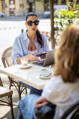 Esmer kadın bir masada oturuyor, açık hava kafesinde dizüstü bilgisayarla çalışıyor. Gündelik kıyafetler ve güneş gözlüğü takan çekici bir kadın..