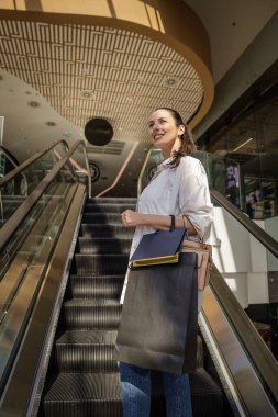 Yerel bir alışveriş merkezinde yürüyen merdivene binen esmer bir kadın. Alışveriş torbaları taşıyor ve aldıkları kıyafetlerle mutlu.. 