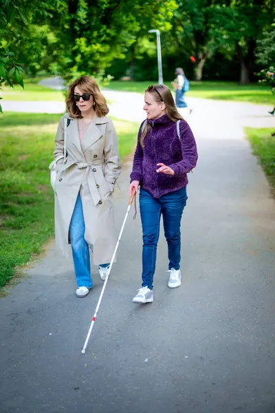 Şehir parkında kadın arkadaşıyla yürüyen görme engelli bir kadın. Görme engelli arkadaşına yardım eden olgun, beyaz bir kadın. Tam uzunluk.  