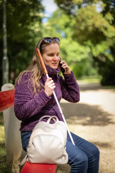 Beyaz baston tutan ve parktaki bankta oturan görme engelli bir kadın. Gülümseyen kadın rahat ceket ve kot pantolon giyiyor..  