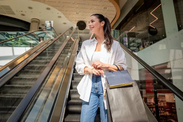 Esmer kadın yerel bir alışveriş merkezinde yürüyen merdivenle aşağı iniyor. Alışveriş torbaları taşıyor ve aldıkları kıyafetlerle mutlu.. 