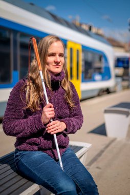 Beyaz bastonlu kör kadın portresi. Şehrin dışındaki tren istasyonunda bankta oturuyor. Treni bekleyen görme engelli bir kadın. Günlük kıyafetler. Tam uzunluk.   