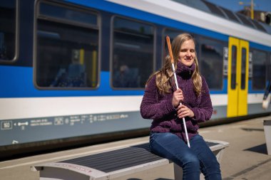 Beyaz bastonlu kör kadın portresi. Şehrin dışındaki tren istasyonunda bankta oturuyor. Treni bekleyen ve telefon kullanan görme engelli bir kadın. Günlük kıyafetler..   