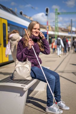 Beyaz bastonlu kör kadın portresi. Şehrin dışındaki tren istasyonunda bankta oturuyor. Treni bekleyen ve telefon kullanan görme engelli bir kadın. Günlük kıyafetler. Tam uzunluk.  