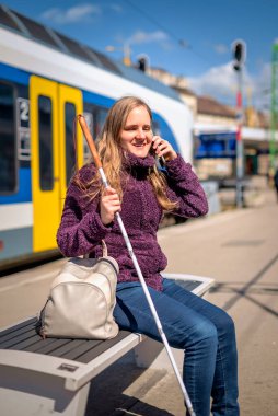 Beyaz bastonlu kör kadın portresi. Şehrin dışındaki tren istasyonunda bankta oturuyor. Treni bekleyen ve telefon kullanan görme engelli bir kadın. Günlük kıyafetler. Tam uzunluk.  
