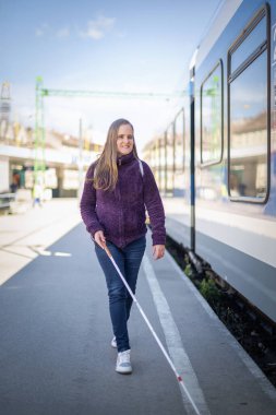 Portrait of blind woman with white cane walking on train station outdoors in city. A vision impaired woman waiting for a train. Casual clothes. Full length.  