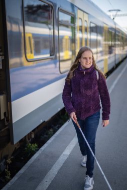 Şehrin dışındaki tren istasyonunda yürüyen beyaz bastonlu kör kadın portresi. Treni bekleyen görme engelli bir kadın. Günlük kıyafetler. Tam uzunluk.  
