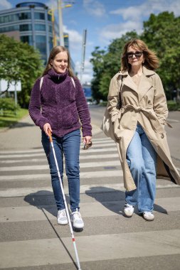 Görme engelli ve işitme engelli bir kadın, kız arkadaşıyla şehirde yürüyor. Görme engelli kadın elinde beyaz bir baston tutuyor ve konuşuyor.. 