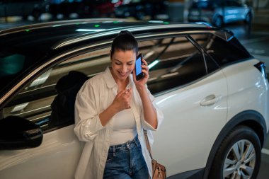 Otoparkta arabasına yaslanan ve telefon kullanan kendine güvenen bir kadın. Esmer kadın, günlük kıyafetler giyiyor ve telefonda konuşuyor..  
