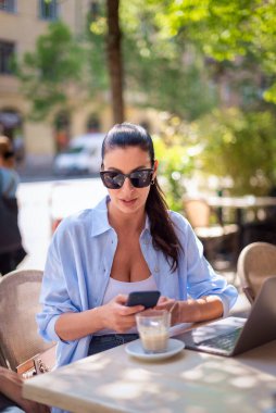 Neşeli bir kadın bir masada oturuyor ve açık bir kafede bilgisayar ve akıllı telefon kullanıyor. Sıradan kıyafetler giyen çekici bir kadın..  