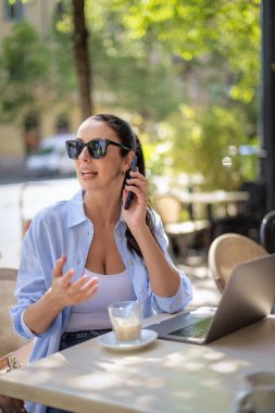 Neşeli bir kadın bir masada oturuyor ve açık bir kafede bilgisayar ve akıllı telefon kullanıyor. Sıradan kıyafetler giyen çekici bir kadın..  