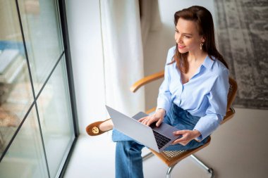 Bilgisayarı kullanan olgun bir kadın. Cam kenarındaki sandalyede oturan esmer saçlı kadın. Merkez Ofis.  