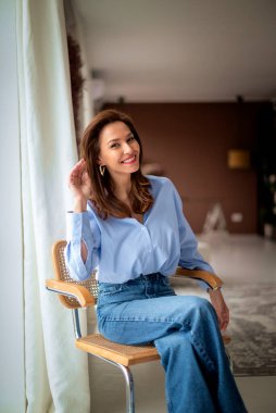 Esmer kadın, pencerenin yanındaki rahat evinde dinleniyor. Sandalyede oturup gündelik giysiler giymek çok çekici..  