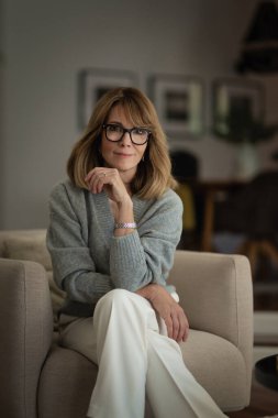 Çekici sarı saçlı kadın modern evinde bir koltukta dinleniyor. Orta yaşlı kadın günlük kıyafetler giyiyor ve gülümsüyor..  