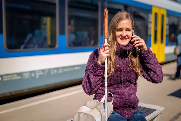 Beyaz bastonlu kör kadın portresi. Şehrin dışındaki tren istasyonunda bankta oturuyor. Treni bekleyen ve telefon kullanan görme engelli bir kadın. Günlük kıyafetler. Tam uzunluk.  