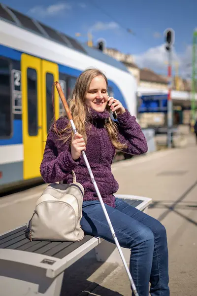 Beyaz bastonlu kör kadın portresi. Şehrin dışındaki tren istasyonunda bankta oturuyor. Treni bekleyen ve telefon kullanan görme engelli bir kadın. Günlük kıyafetler. Tam uzunluk.  