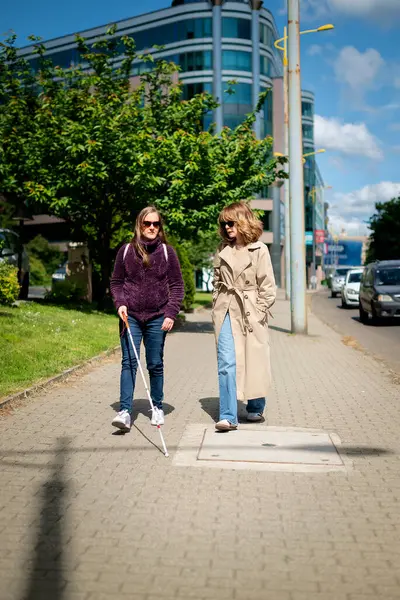 Görme engelli ve işitme engelli bir kadın, kız arkadaşıyla şehirde yürüyor. Görme engelli kadın elinde beyaz bir baston tutuyor ve konuşuyor.. 