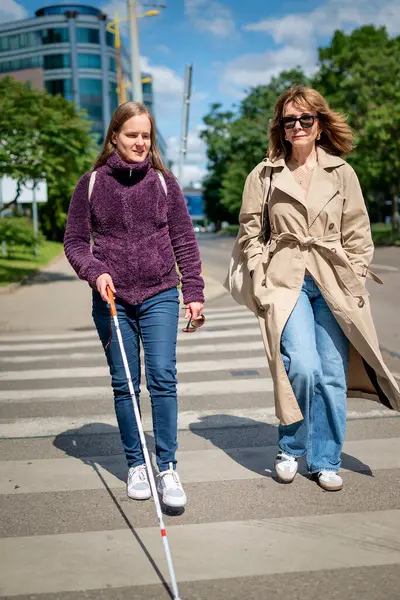 Görme engelli ve işitme engelli bir kadın, kız arkadaşıyla şehirde yürüyor. Görme engelli kadın elinde beyaz bir baston tutuyor ve konuşuyor.. 