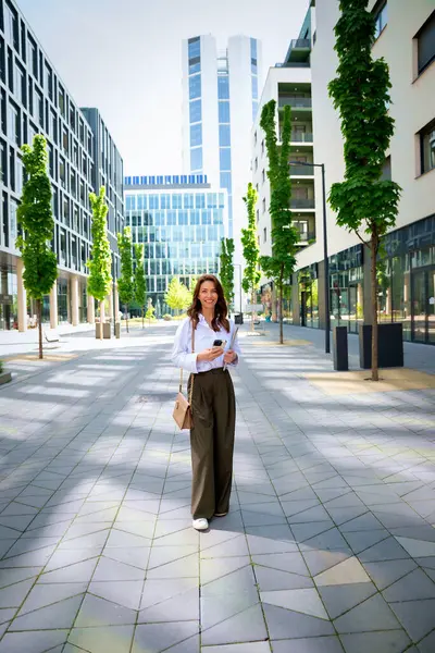 İş yerindeki ofise yürüyen kendine güvenen bir iş kadını. Elinde akıllı telefon ve bilgisayar dizüstü bilgisayarı tutan profesyonel bir kadın. Esmer, iş kıyafeti giyen bir kadın.. 
