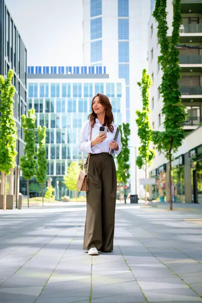 İş yerindeki ofise yürüyen kendine güvenen bir iş kadını. Elinde akıllı telefon ve bilgisayar dizüstü bilgisayarı tutan profesyonel bir kadın. Esmer, iş kıyafeti giyen bir kadın.. 